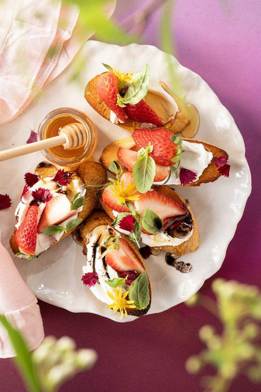 Strawberry and Goat Cheese Crostini on a white plate with a pink napkin and one 3 ounce jar of honey and covered in strawberries and edible garnishes.
